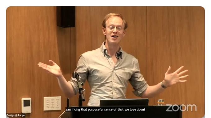 Andy Matuschak standing at a podium mid-gesture, arms open, in a wood-panelled lecture room