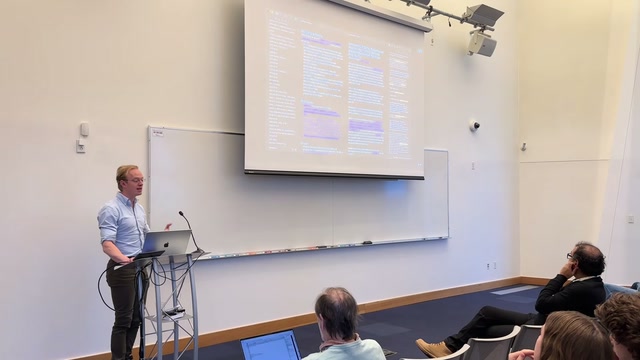 A presenter at a podium gestures as he delivers a lecture to a seated audience, with a software interface displayed on a large projector screen above him.